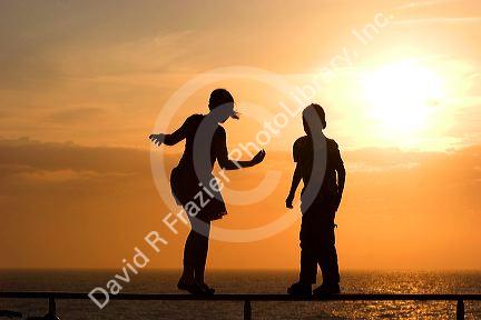 Silhouette of children walking on a railing at sunset by the sea.