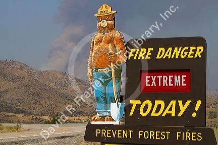 Smokey Bear extreme fire danger sign with a plume of smoke from a forest fire in the back ground in Boise County, Idaho, USA.
