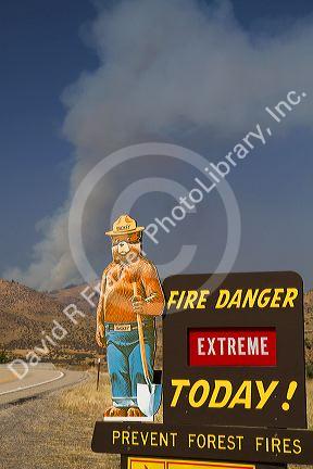 Smokey Bear extreme fire danger sign with a plume of smoke from a forest fire in the back ground in Boise County, Idaho, USA.