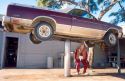 A male adult auto mechanic stands under a car upon a hydraulic lift.
